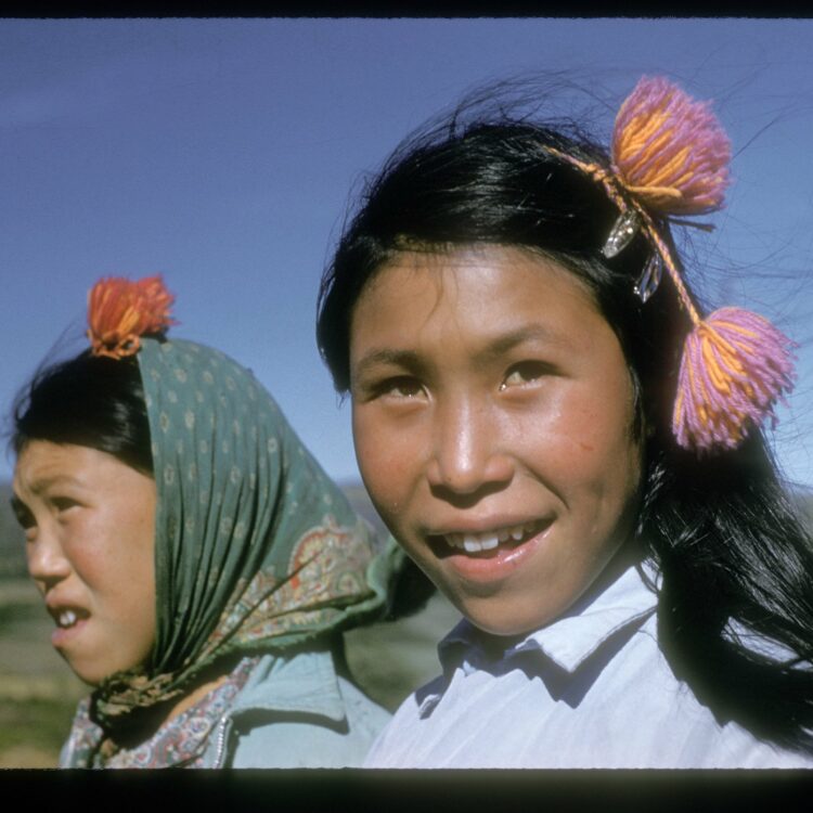 A close-up shot of two young girls standing in an exterior location. The girl on the right is smiling slightly, and staring off just behind the camera. The girl on the right is behind the first, wearing a green head covering and staring off camera to the left.
