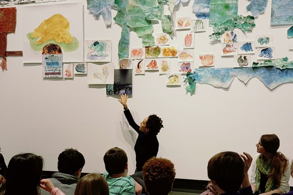 Artist Lucie Chan seated in front of wall with large art papers hung on the wall with children gathered around listening