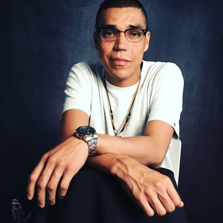 Studio photograph of Michael Roderick Keshane, a young Cree/Anishinaabe man in a white t-shirt with a flashy watch and gold chains, comfortably at ease.