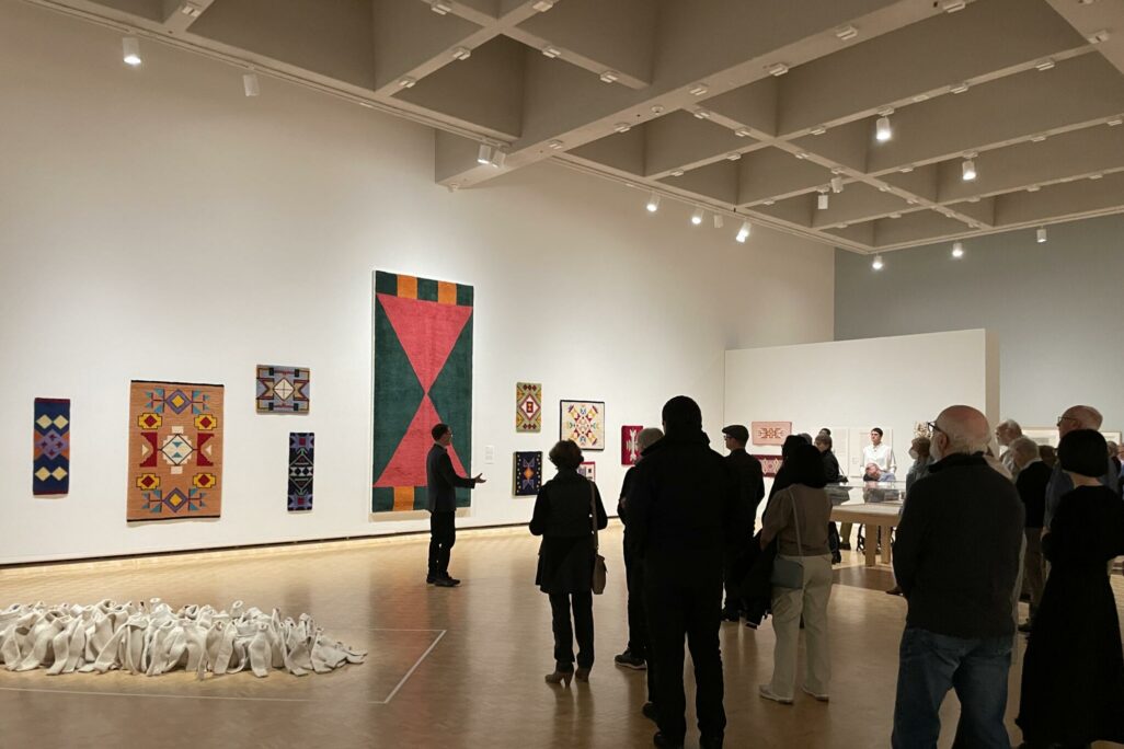 A group of visitors standing in front of large rugs displayed on the wall as a curator describes the work to them.
