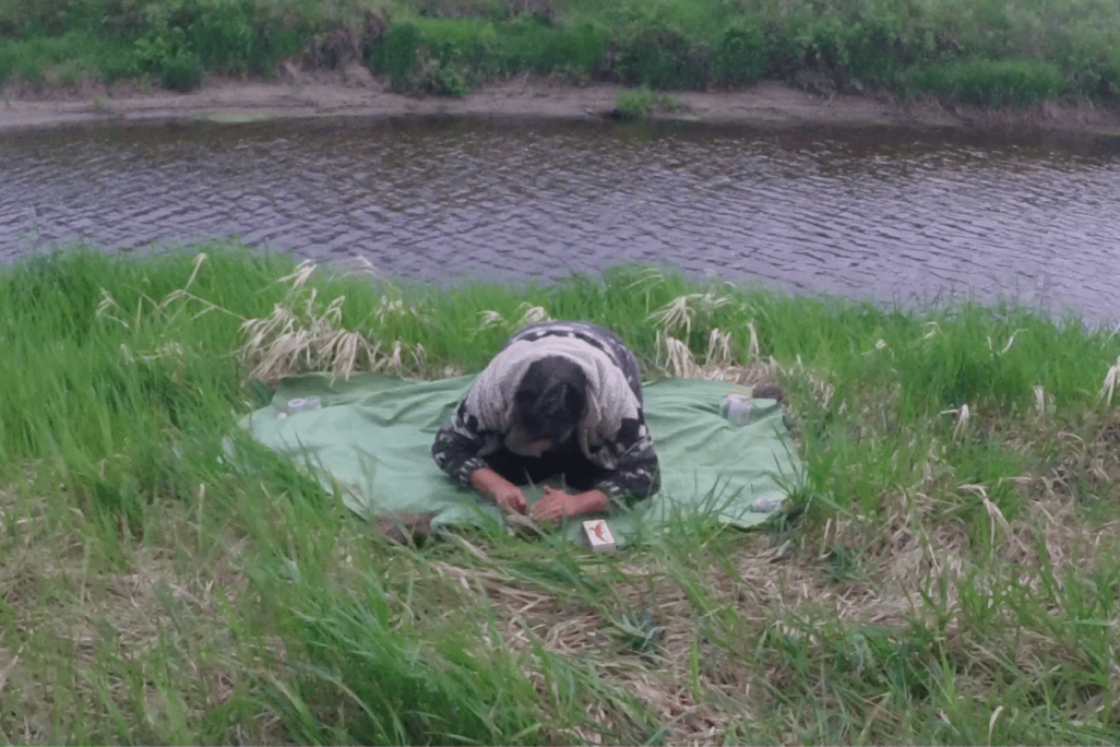 A person kneeling on the grass near a river. The person is facing away from the water kneeling on a blanket with their head to the ground in a deep bow.