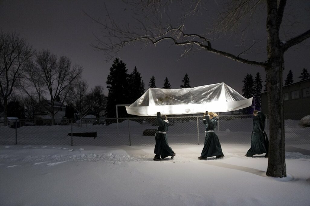 Three figures march through the winter snow carrying a transparent canoe shaped structure that glows from the inside.