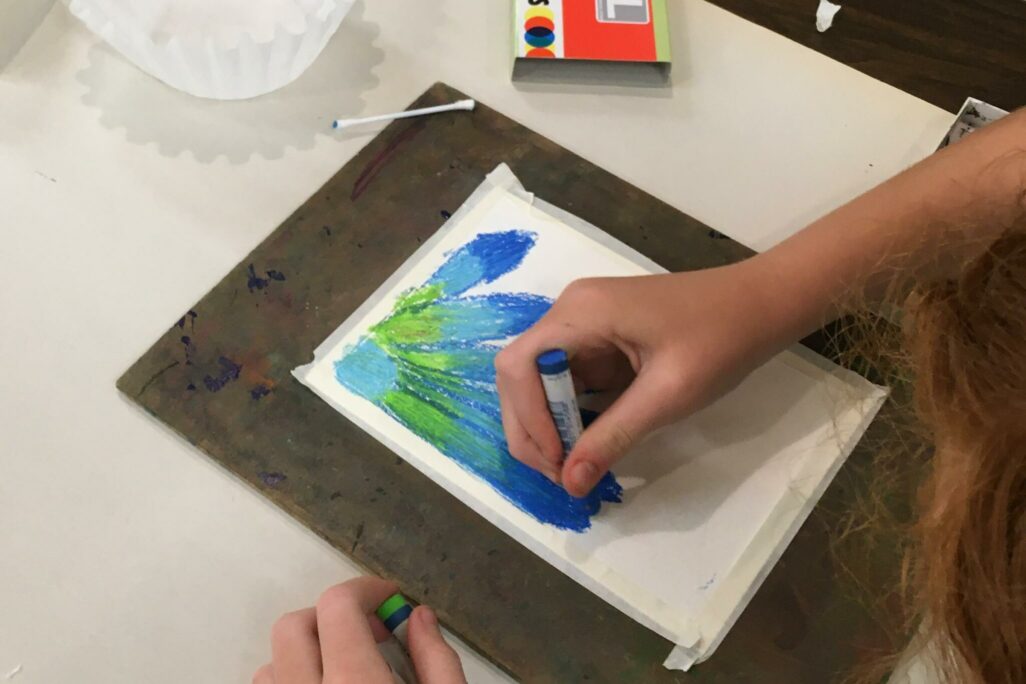 A child's hands drawing a flower in shades of blue and green with pastel.