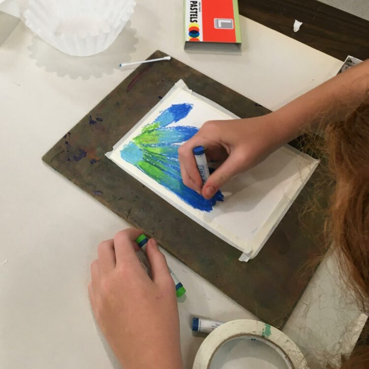 A child's hands drawing a flower in shades of blue and green with pastel.