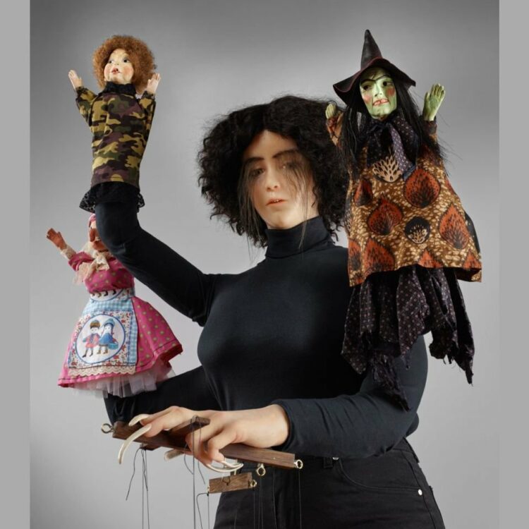A wax figure of a lady in all black with four arms, each arm is controlling a small puppet.